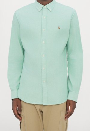 Mężczyzna w jasnooliwkowej koszuli zapinanej na guziki z kołnierzykiem i małym haftowanym logo na piersi oraz beżowych spodniach typu cargo.