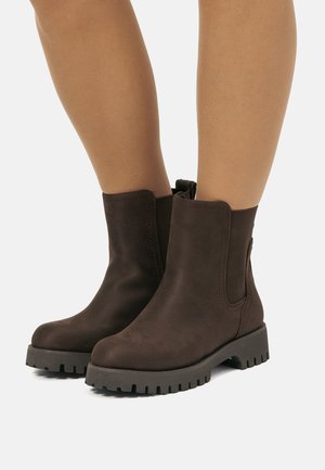 Un par de botas de tobillo de ante marrón oscuro con suelas de goma resistentes, usadas sobre piernas desnudas contra un fondo claro y liso.