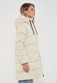 Cappotto imbottito beige con cappuccio, dotato di chiusura a zip e coulisse, ampie sezioni trapuntate e dettagli neri attorno al collo.