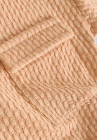 Pfirsichfarbener Stoff mit einer strukturierten, waffleartigen Musterung, versehen mit einem umgeschlagenen Saum und einem kleinen Taschendetail. Weiches, leichtes Material.