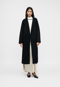 Μακρύ μαύρο παλτό με μεγάλα πέτα, κατασκευασμένο από υλικό με μείγμα μαλλιού. Διαθέτει ίσια γραμμή και ανοιχτό μπροστινό σχέδιο, φορεμένο πάνω από κρεμ φόρεμα και τακούνια.