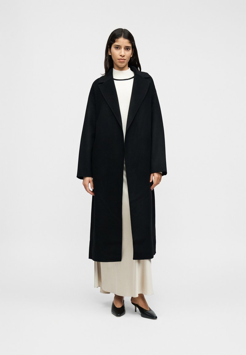 Μακρύ μαύρο παλτό με μεγάλα πέτα, κατασκευασμένο από υλικό με μείγμα μαλλιού. Διαθέτει ίσια γραμμή και ανοιχτό μπροστινό σχέδιο, φορεμένο πάνω από κρεμ φόρεμα και τακούνια.