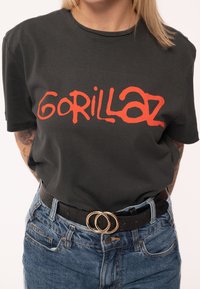 Black cotton t-shirt with red "Gorillaz" graphic print. Pairing with high-waisted blue denim jeans and a black belt with double gold rings.