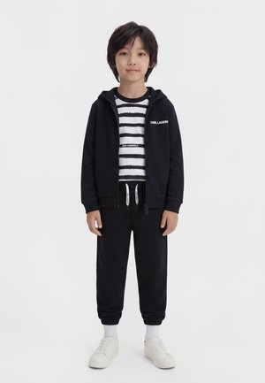 Bambino in piedi, indossa una felpa nera con zip, una maglietta a righe nere e bianche, pantaloni da jogging neri, calzini bianchi e sneakers bianche su uno sfondo semplice.