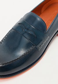 Σκούρο μπλε δερμάτινη λοφάκια με λεία επιφάνεια, που διαθέτει διακοσμητική ραμμένη ζώνη και μια διακριτικά ανυψωμένη λογότυπη λεπτομέρεια. Αντίθετο καφέ εσωτερικό.