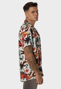 Camisa de manga corta con cuello en tejido blanco, que presenta un colorido patrón tropical con palmeras, flores y tablas de surf.