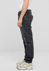 Jeans de mezclilla negra con un corte relajado, que presentan detalles desgastados y costuras visibles. Combinados con zapatillas beige.