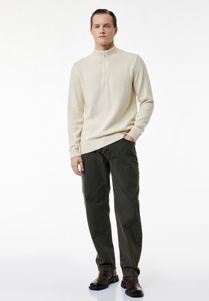 Homme debout les mains dans les poches, portant un pull crème à fermeture éclair quart, pantalon vert foncé et bottes marron foncé sur fond blanc.