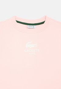 Sweatshirt rosa claro com gola redonda, gola interior verde e logótipo branco de crocodilo com o texto "LACOSTE PARIS". Textura suave.