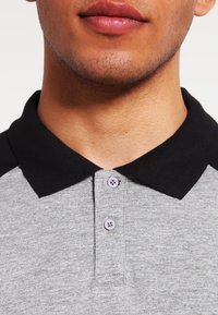Polo gris avec un col noir, trois boutons violets et un tissu texturé. Focus sur les détails du col et des boutons.