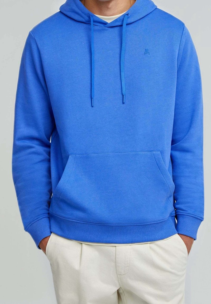 Homme portant un sweat à capuche bleu vif avec poche avant et cordons, associé à un pantalon beige clair, mains dans les poches, sur fond uni.