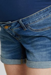 Calções de maternidade em denim com lavagem média azul, apresentando fecho de botão, bainhas dobradas e bolsos frontais, projetados para conforto.