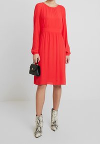 Robe rouge à manches longues avec un col et une taille froncés. Associée à un petit sac à main noir et des bottines blanches à imprimé serpent.