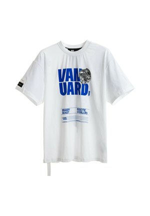 Joven de pie vistiendo una camiseta blanca con texto y gráfico azul, pantalones cortos de bloques de color en blanco, rojo y azul, calcetines blancos y zapatos beige.