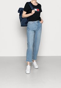 Svart bomull t-shirt med logotyp, ljusblå denimjeans med fransade fållar, och vita sneakers. Mörkblå ryggsäck hålls i ena handen.