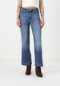 Jeans de mezclilla azul de tiro alto y pernera acampanada con detalles de botones metálicos a los lados y un dobladillo sin terminar. Combinados con botas de tacón de ante marrón.
