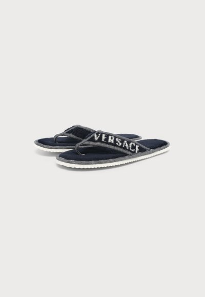 Tongs Versace bleu marine avec semelle blanche et logo sur les lanières, exposées sur un fond gris clair.