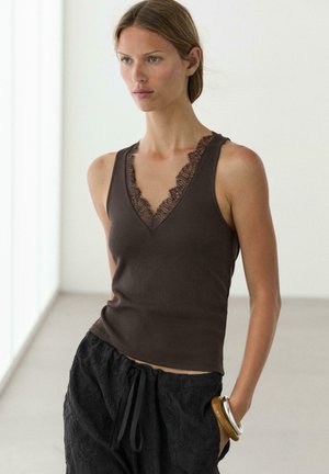 Frau trägt ein ärmelloses dunkelbraunes Oberteil mit Spitzenbesatz und schwarze, strukturierte Hose, steht in einem hellen, minimalistischen Raum.