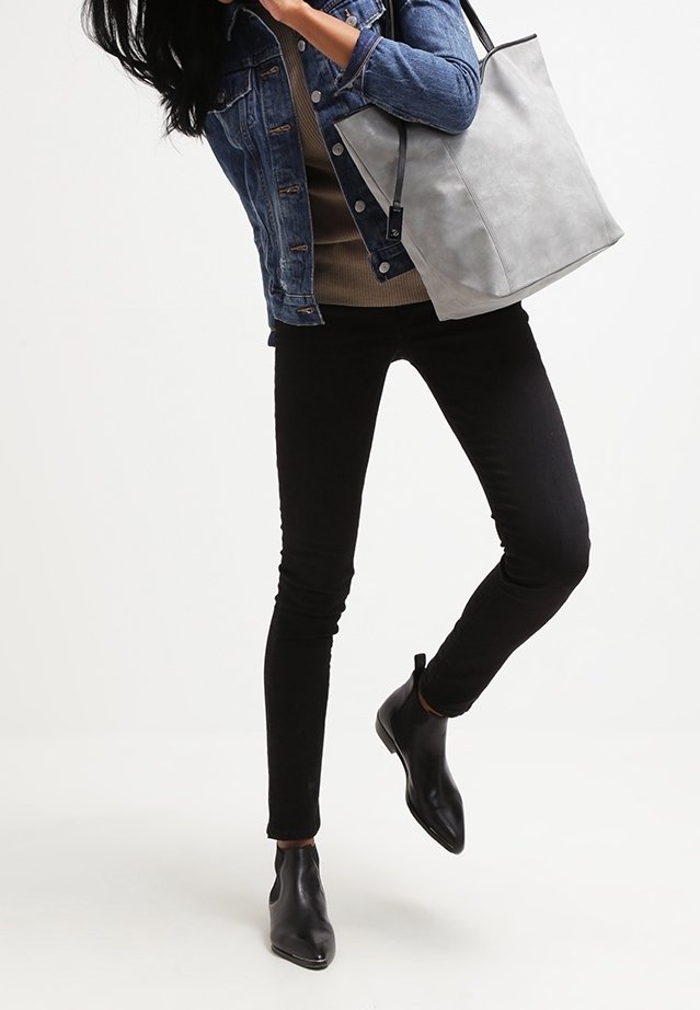 Femme portant un pantalon slim noir, des bottines noires, une veste en jean, avec un grand sac cabas gris clair à l'épaule.