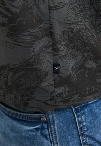 Černé tričko s jemným abstraktním vzorem, krátkými rukávy a malou látkovou štítkovou etiketou na lemu, ve spojení s modrými džínami.