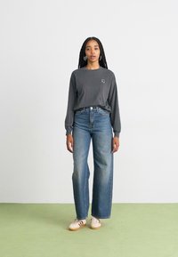 Gray long-sleeve shirt with a small logo, paired with high-waisted wide-leg blue jeans and cream shoes with dark stripes. Plain background.