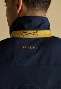 Vīrietis valkā tumši zilu jaku ar dzelteno apkakles maliņu, izšūtām krustām polo nūjām un tekstu "JOULES" aizmugurē netālu no kakla.