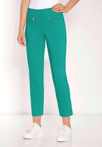 Pantalons teal à taille élastique avec deux poches avant zippées, coupe slim, longueur cheville, associés à des baskets blanches sur un fond neutre.