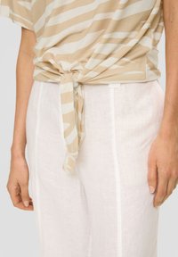 Beige und weiß gestreiftes Oberteil mit einem Knoten in der Taille, kombiniert mit lockeren weißen Leinenhosen mit Seitennaht und texturierter Oberfläche.