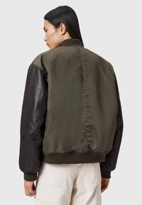 Personne avec de longs cheveux noirs portant une veste bomber verte olive avec des manches en cuir noir et un pantalon beige, de dos par rapport à l'appareil photo.