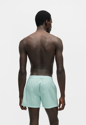 Mężczyzna o ciemnej karnacji i krótkich kręconych włosach widziany od tyłu, ubrany w jasnoniebieskie spodenki na jednolitym białym tle.