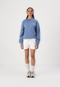 Sweatshirt bleu clair avec logo, short blanc et baskets roses. Manches longues, coupe décontractée, en coton. Chaussettes blanches côtelées.