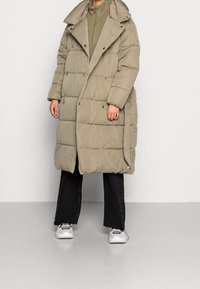 Abrigo acolchado beige con un cuello alto, que presenta un diseño acolchado, cierres de botón y bolsillos laterales, combinado con pantalones negros y zapatillas.