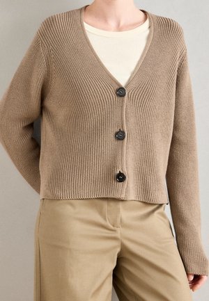 Cardigan beige en tricot côtelé avec trois boutons noirs, porté sur un top crème et associé à un pantalon beige.