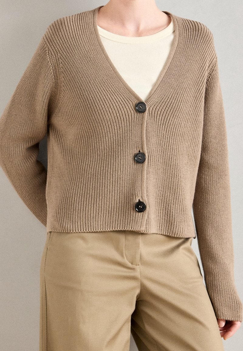 Cardigan beige en tricot côtelé avec trois boutons noirs, porté sur un top crème et associé à un pantalon beige.