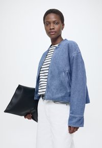 Ženska s kratkimi lasmi, oblečena v modro denim jakno, črtasto majico in bele hlače, ki drži veliko črno torbico na enobarvni podlagi.