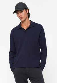 Man wearing a navy long-sleeve polo shirt, black pants, and a black baseball cap, posing with hands in pockets against white background.