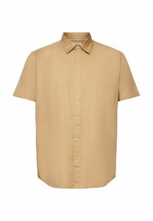 Person trägt beige kurzärmliges Hemd mit Knöpfen, beige Cargohosen, weiße Turnschuhe und einen orangefarbenen Pullover um die Taille gebunden.