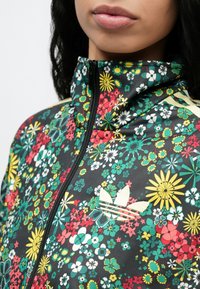 Frau trägt eine gestreifte Blumenjacke mit gelben, roten, weißen und grünen Blumen und einem weißen Adidas-Logo auf der Brust.