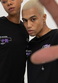 Black cotton t-shirts with purple and white graphic prints, featuring text and symbols. Close-up of two individuals, partially obscured by a hand.