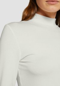 Haut à manches longues blanc avec un col montant, fabriqué en tissu lisse. Présente une coupe ajustée et un minimum de détails de couture.