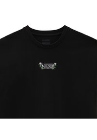 Camiseta negra de algodón con cuello redondo y mangas cortas. Presenta un logo bordado en blanco y verde de "VANS" rodeado de diseños de hojas de palma.