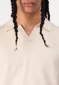 Krämfärgad polo skjorta med V-hals, mjuk textur och kort, vikkant; har en liten fransad detalj nära halslinjen.