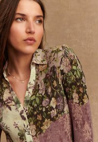 Blouse met bloemenpatroon en geplooide textuur, met tinten groen, paars en crème. Knoopsluiting bij de kraag en delicate bloemaccenten.