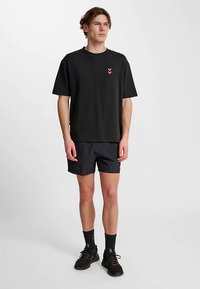 Svart bomullst-shirt med en liten röd chevron-logotyp, kombinerad med mörka shorts, svarta strumpor och träningsskor, stående på en enfärgad bakgrund.