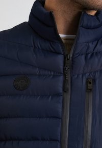 Marineblauer Pufferjacke mit einem hohen Kragen. Verfügt über horizontale Nähte, ein rundes Logo-Patch und zwei mit Reißverschluss verschlossene Taschen. Glatte Textur.