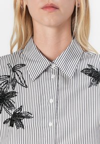 Kvinde iført en sort og hvid vertically striped skjorte med sorte perleblomsterbroderier på skuldrene.