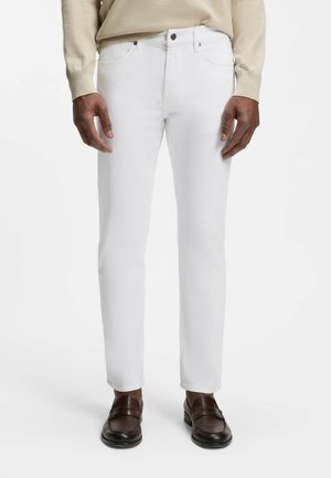 Mand iført slim-fit hvide jeans, beige sweater og mørkebrune læder loafers, står mod en hvid baggrund.