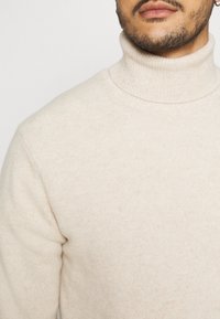 Suéter de cuello alto beige, tejido de punto suave, silueta ajustada, textura acanalada en el cuello y los puños, diseño minimalista sin patrones ni acentos.