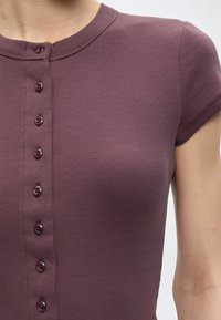 Gros plan d’une personne portant un haut violet côtelé à manches courtes, boutonné, avec un col rond.