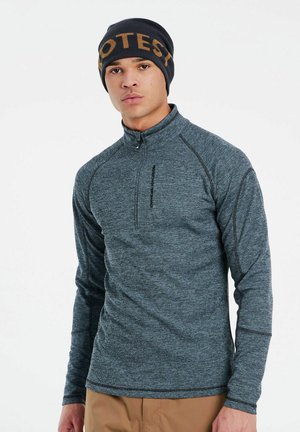 Grijze fleece met rits en hoge kraag en lange mouwen. Heeft contrasterende stiksels en een logo op de borst. Draagt een zwarte beanie.